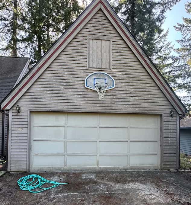 Grimy tan lap-siding garage with stained doors, basketball hoop and a stained concrete apron before washing.