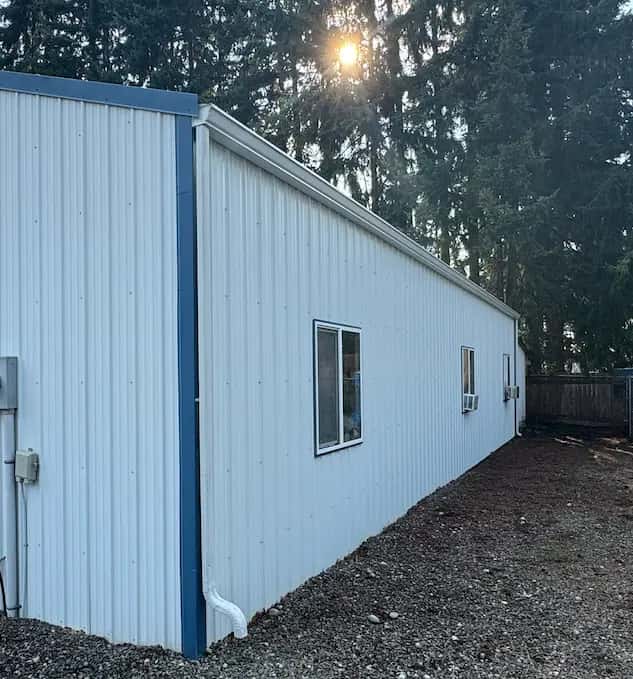Long white metal-sided commercial building with blue corner trim, clean after exterior washing, with evergreens behind.