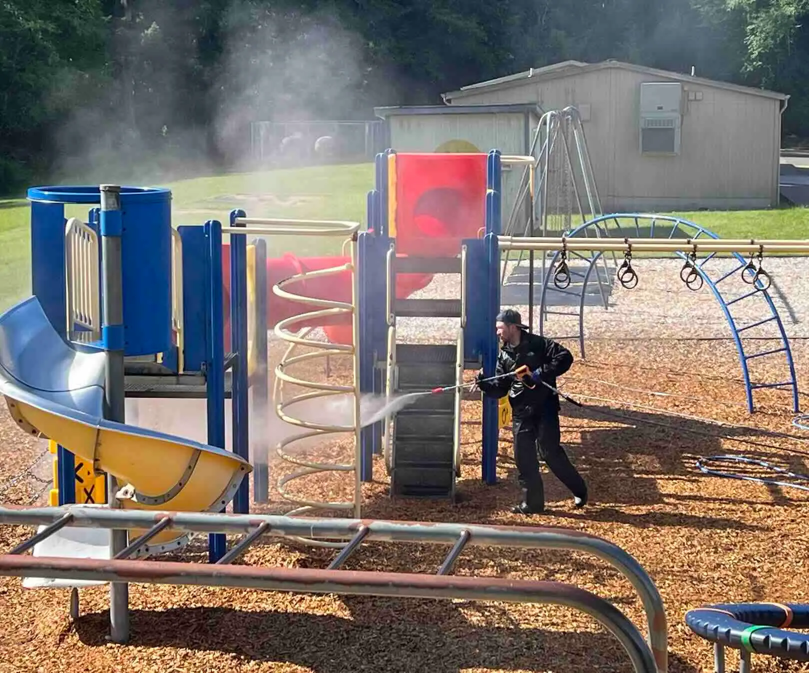Technician pressure washing a blue, red and yellow playground structure with slides on a wood-chip schoolyard.