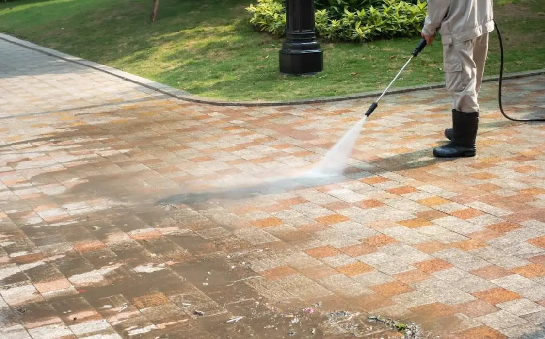 Worker in rubber boots pressure washing a red and tan brick paver walkway, dirty water running off the cleaned pavers.
