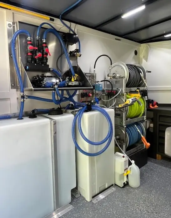 Inside a soft wash trailer: white chemical tanks, blue hoses, a pump manifold and three stacked hose reels.
