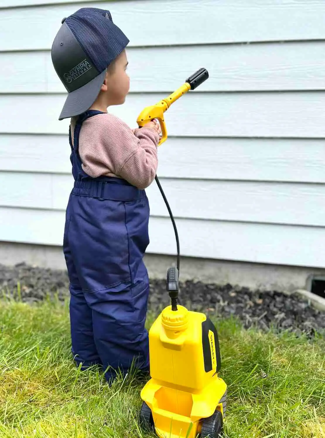 Toddler in an Olympia Softwash cap and navy overalls aiming a yellow toy pressure washer at light blue lap siding.
